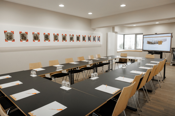 Seminarraum mit U-förmig angeordneten Tischen, ausgestattet mit Notizblöcken, Stiften und Gläsern, große Fensterfront im Hintergrund.