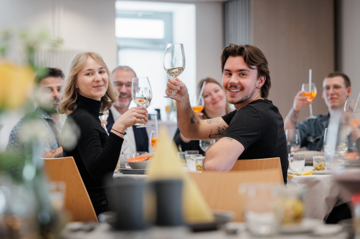Mehrere Personen sitzen gemeinsam an einem Tisch, stoßen mit Getränken an und lachen in einem Restaurant.