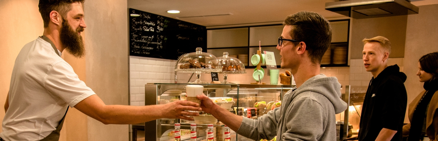 Mann überreicht Kunde hinter Tresen ein Getränk.