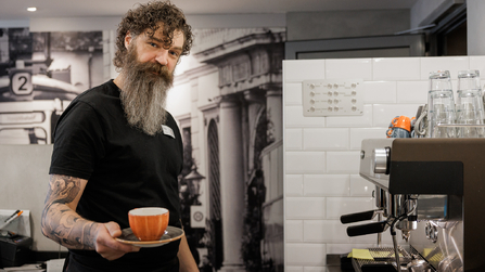Barista mit Bart hält eine Tasse Kaffee vor einer Espressomaschine.